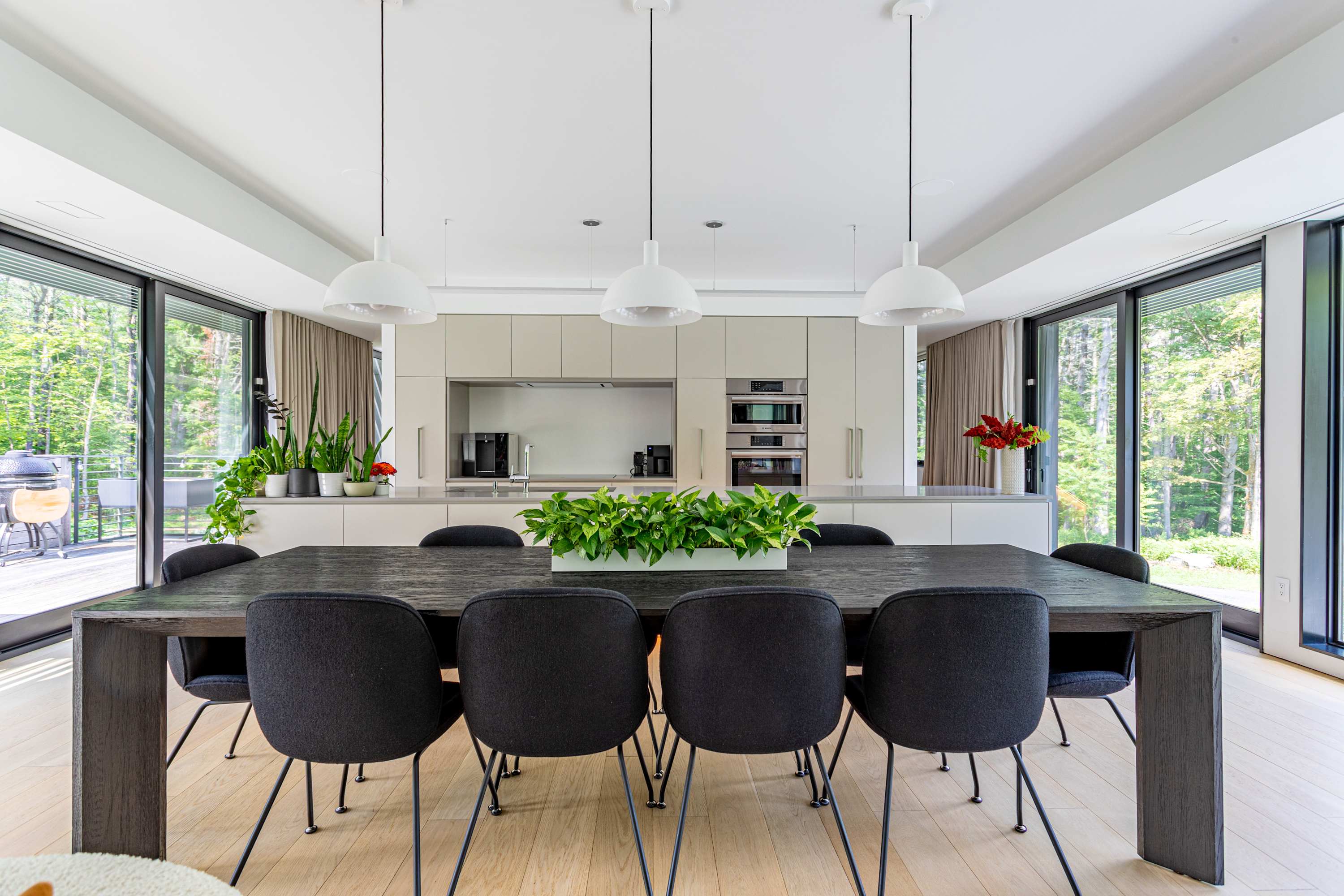 Modern dining area featuring a long dark wood table with black upholstered chairs, illuminated by three white pendant lights, adjacent to a minimalist kitchen and large windows offering forest views.