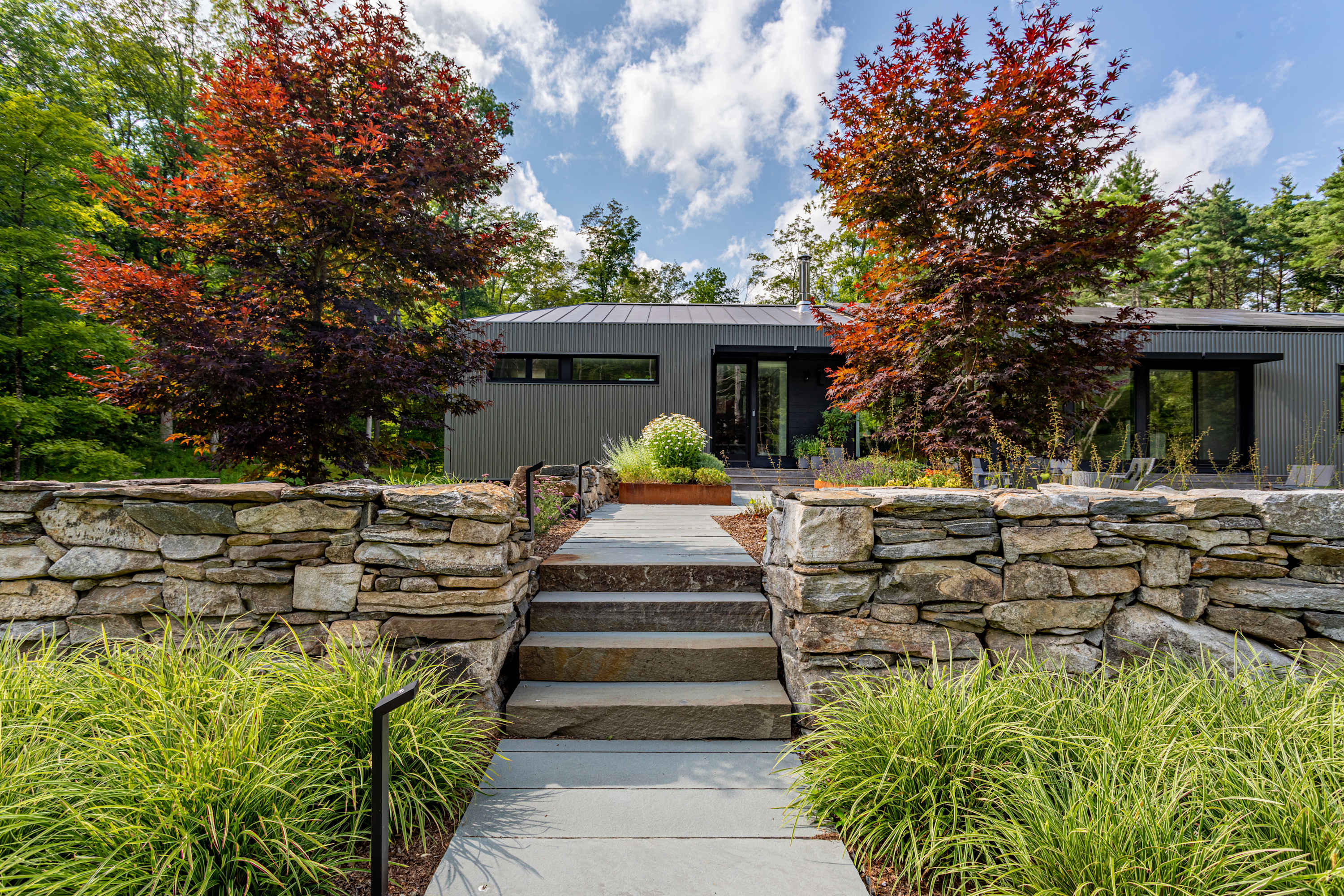 The front exterior of the contemporary house, approached by stone steps and a walkway, flanked by stone walls and vibrant red-leafed trees. The house features dark siding and large windows, blending with the natural landscape.