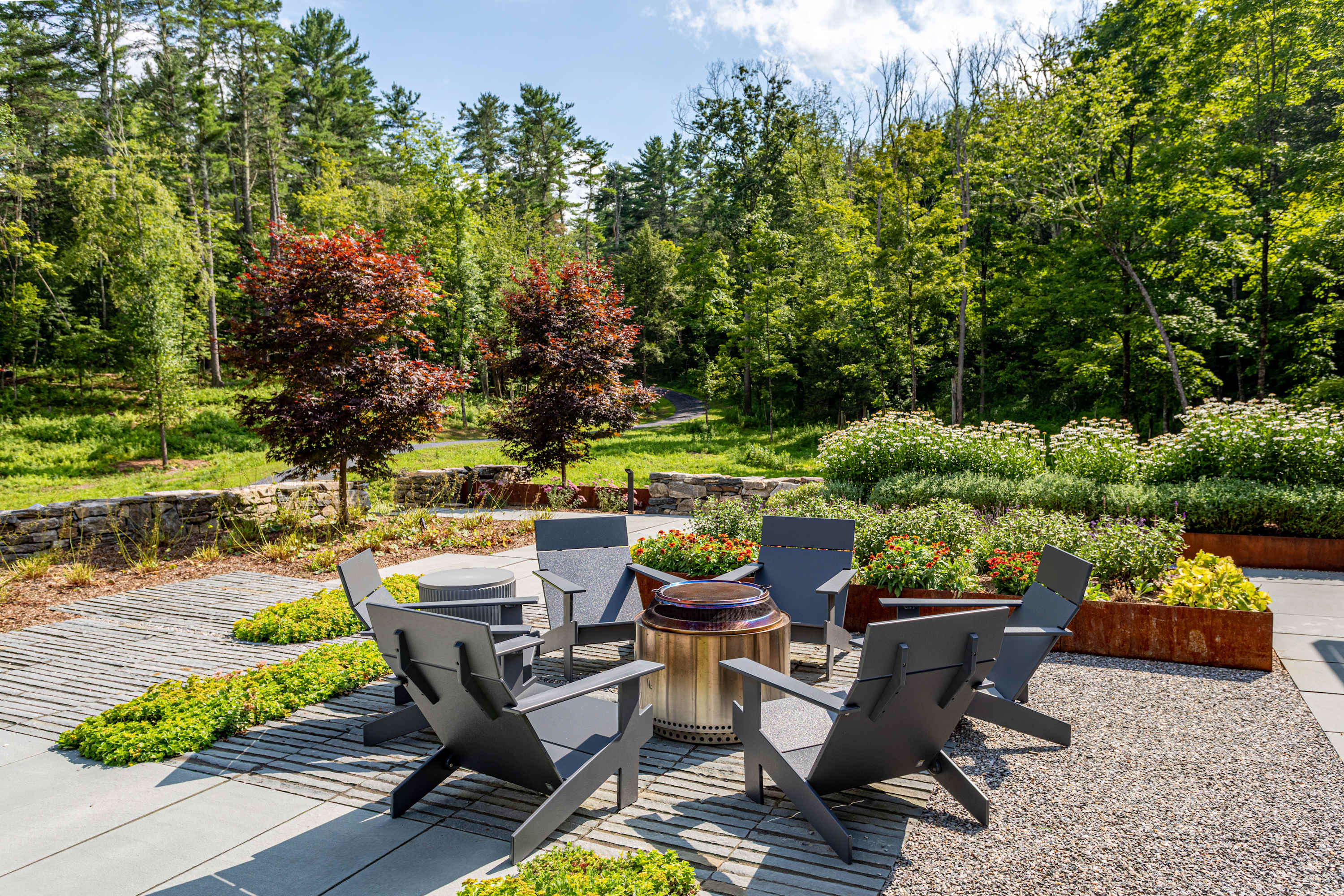 An inviting outdoor seating area with a circular arrangement of modern grey chairs around a central fire pit. The patio is paved with a mix of concrete and gravel, surrounded by landscaped garden beds and mature trees.