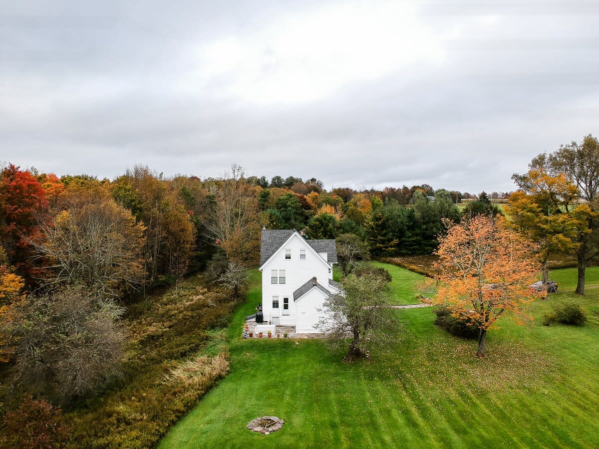 A wide aerial shot showcasing a white house with a dark roof nestled amidst a vast expanse of green lawn and dense autumn forest with colorful foliage. The property appears secluded and surrounded by nature under an overcast sky.