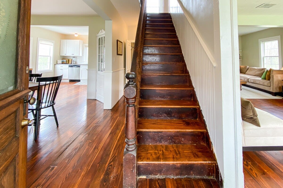 A rustic wooden staircase with dark stained treads and white risers leads upstairs, visible from a foyer with dark hardwood floors. The living room and dining room are visible through doorways on either side, showcasing the home's flow.