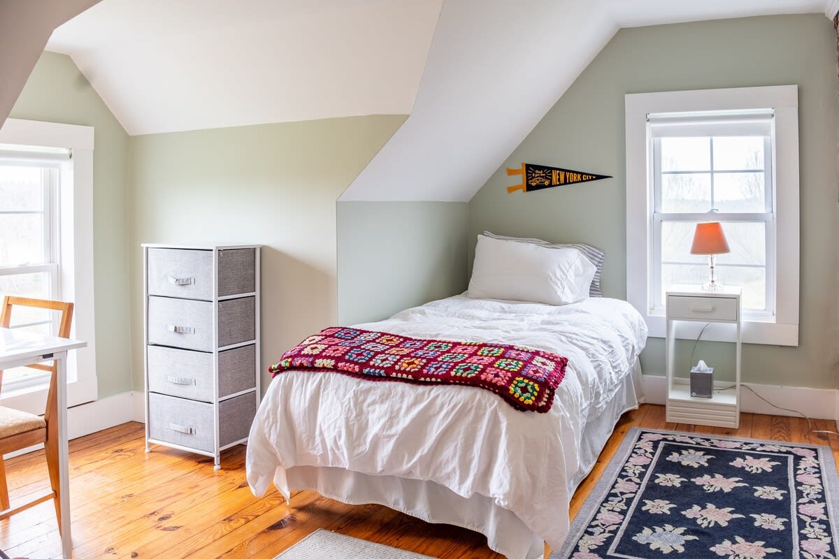 This cozy bedroom features a single bed with a white duvet and a colorful crocheted blanket, set against a light green wall with a sloped white ceiling. A small nightstand with a lamp is next to the bed, and the room has warm hardwood floors and a patterned rug.
