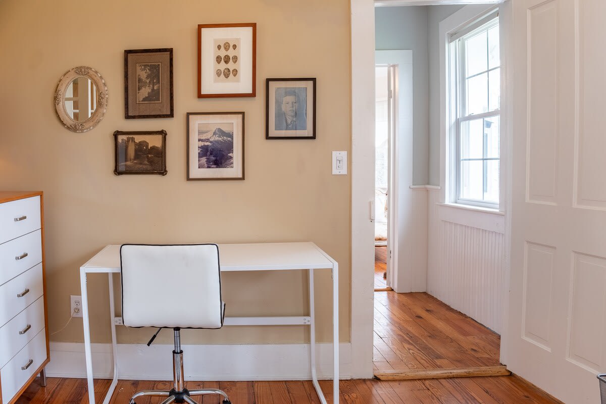 A dedicated office space with light brown walls and hardwood floors, featuring a white desk and a black and white office chair. A gallery wall with various framed artworks adds character to the space.