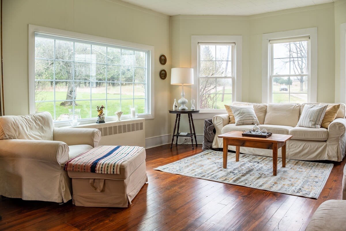A cozy living room features light green walls, dark hardwood floors, and a large window overlooking a grassy outdoor area. A cream-colored armchair and ottoman sit opposite a matching sofa, centered around a wooden coffee table and an area rug.