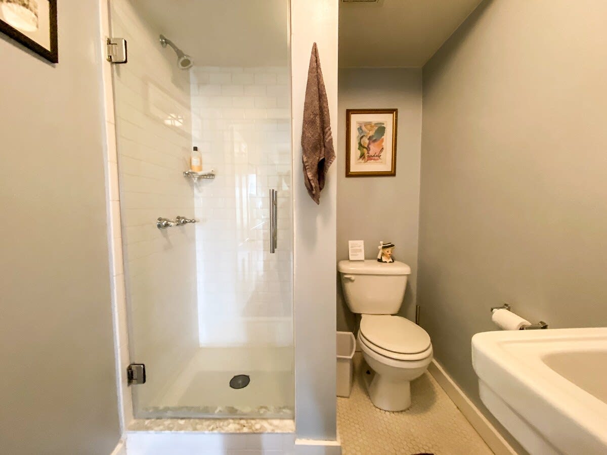 A modern bathroom with light gray walls and a white hexagonal tile floor. It features a glass-enclosed shower with subway tiles, a toilet, and a white pedestal sink, with a framed picture adding a touch of decor.