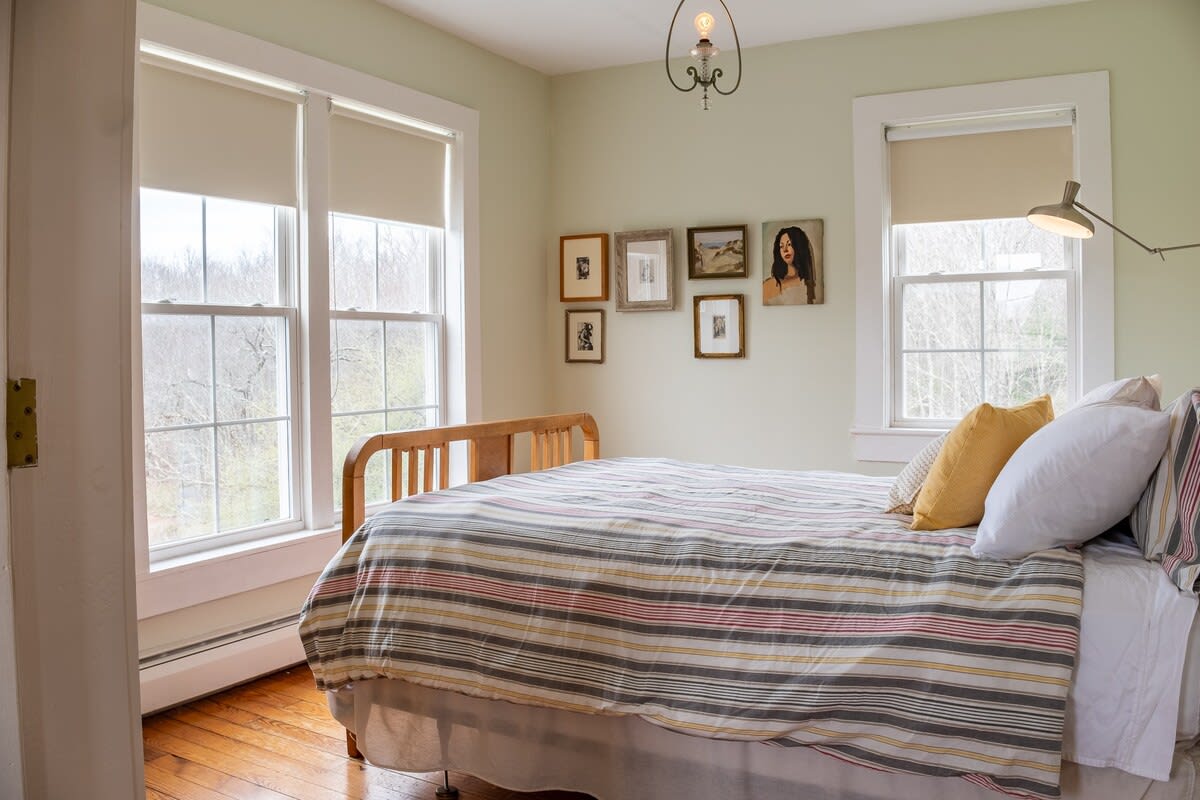 A bright bedroom with light green walls and hardwood floors, featuring a bed with a striped duvet and multiple large windows. Framed artwork adorns the wall above the bed, adding a personal touch.