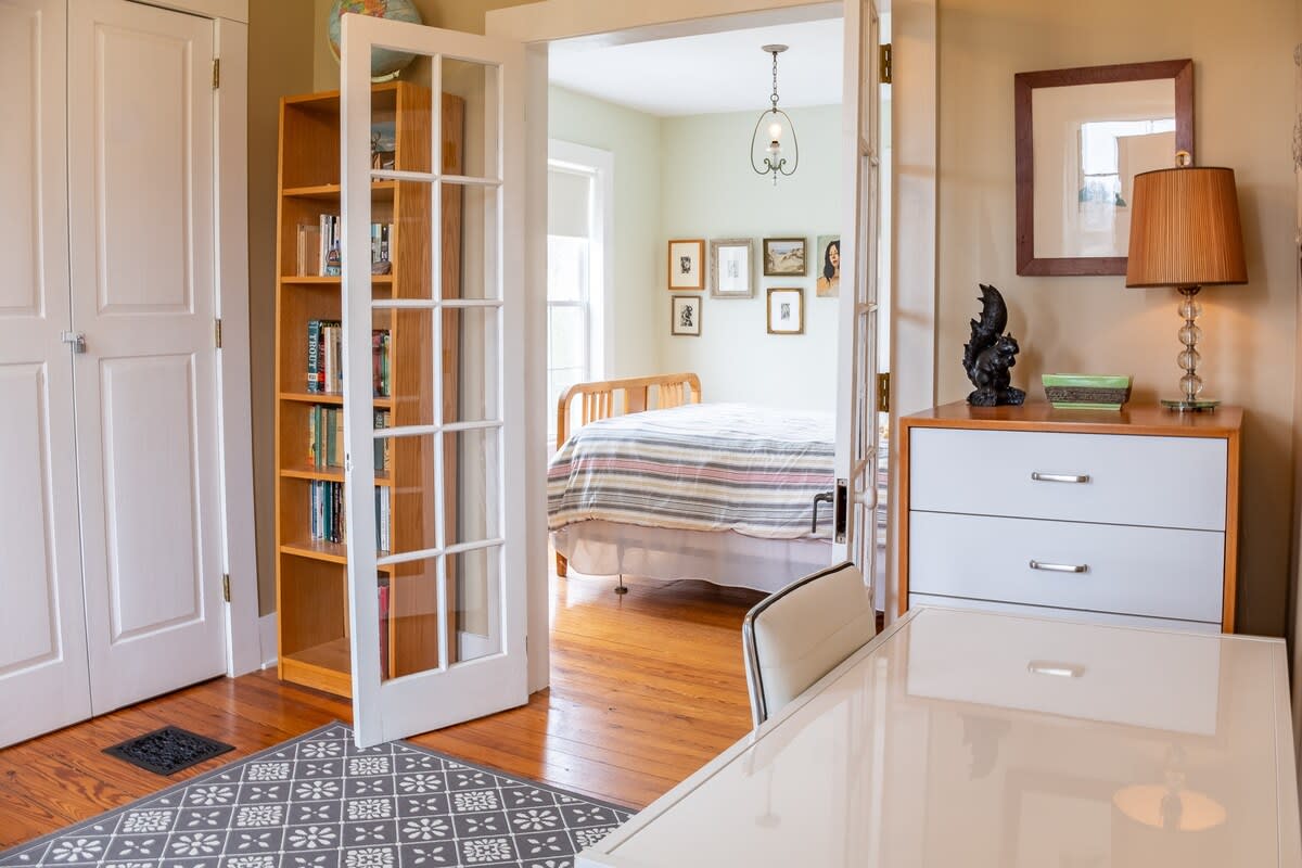 An office space with light brown walls and hardwood floors, featuring a white desk and chair, and a bookshelf. French doors open into a bedroom with a bed and multiple windows, creating a bright and functional area.