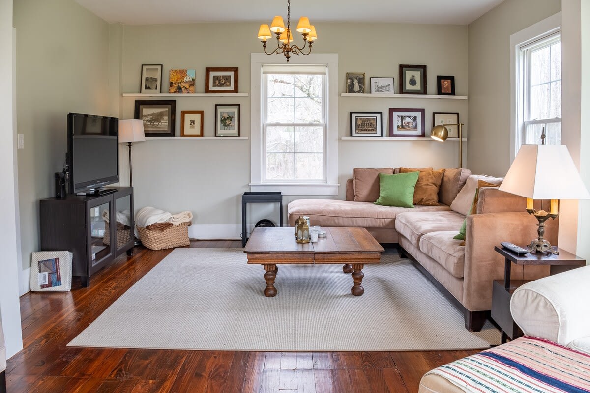 Cozy living room with dark hardwood floors and light green walls, featuring a large L-shaped sectional sofa in a neutral tone, a rustic wooden coffee table, and a light-colored area rug. Shelves with framed pictures adorn the wall.