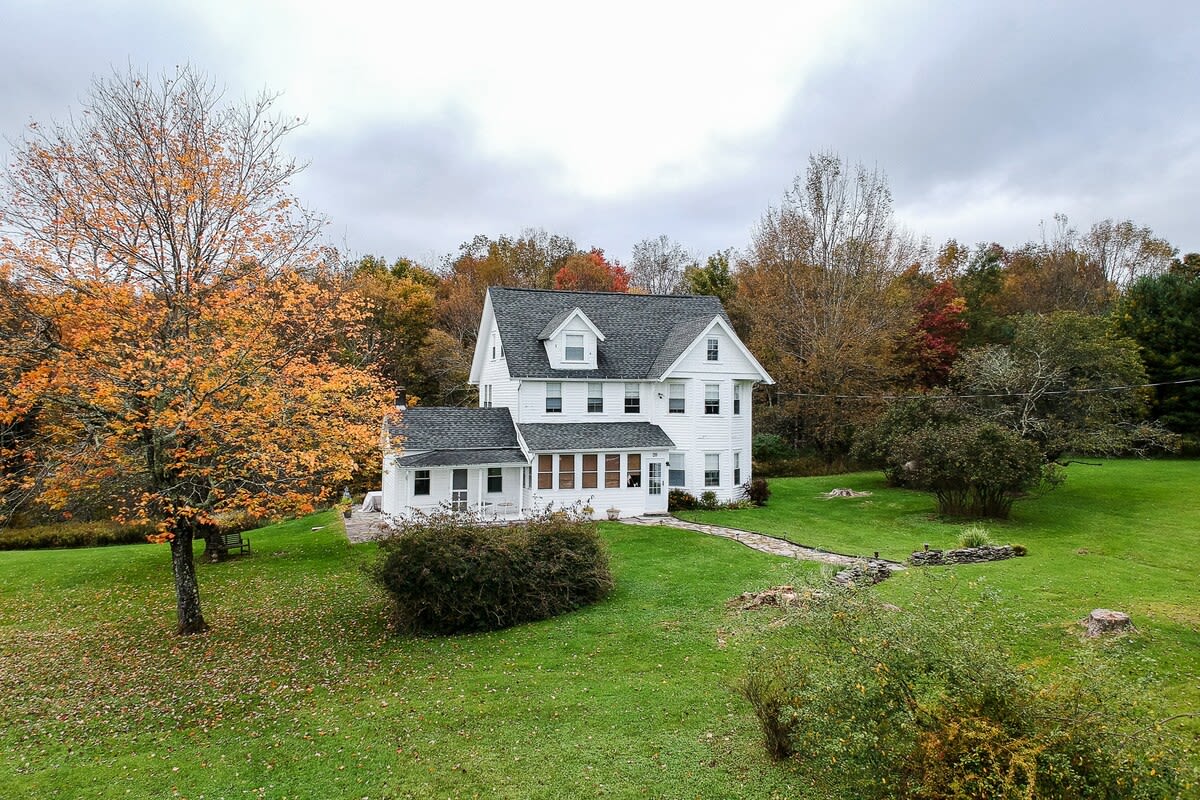 An aerial view of a white two-story house with a dark gray roof, surrounded by a lush green lawn and vibrant autumn trees with orange and red foliage under a cloudy sky. A stone path leads to the front entrance.