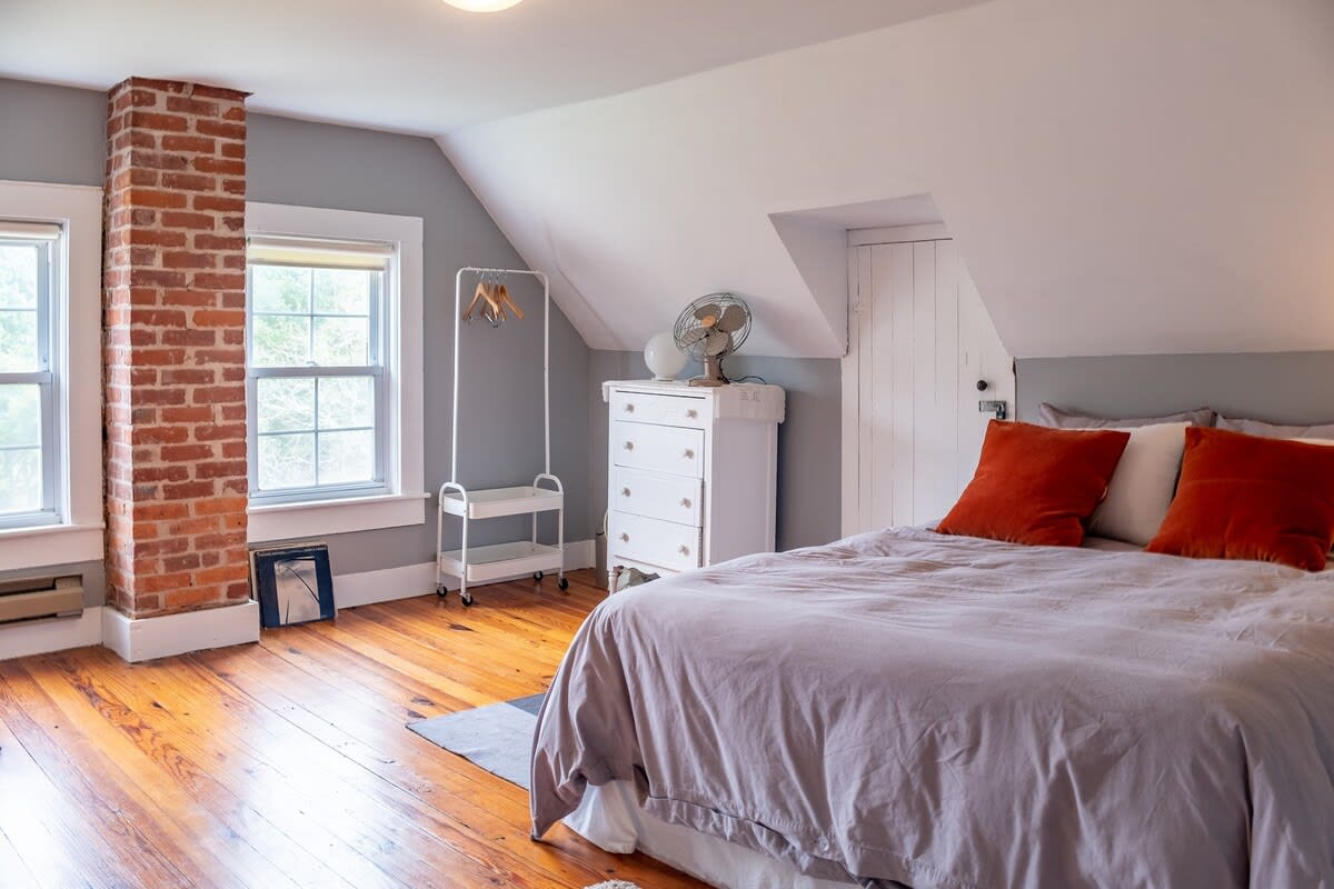 This bedroom features a large bed with a gray duvet and orange pillows, set against a light gray wall with a sloped white ceiling. A prominent exposed brick chimney column stands next to two windows, and the room has shiny hardwood floors.