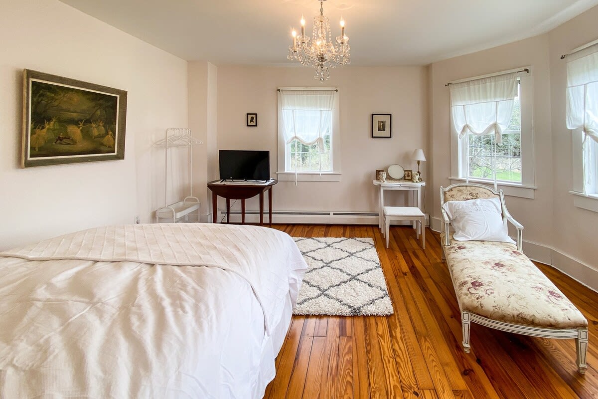 A spacious bedroom with light walls and polished hardwood floors, featuring a white-covered bed and a decorative chaise lounge. Multiple windows provide ample natural light, and a crystal chandelier hangs from the ceiling.