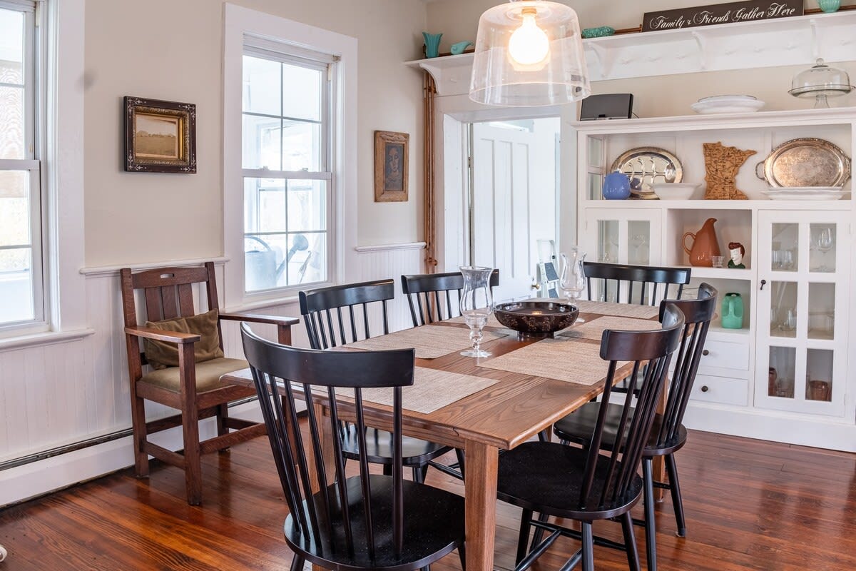Inviting dining area with a wooden table and black spindle-back chairs, set on dark hardwood floors. A white built-in hutch displays dishware, and large windows provide natural light, complementing the clear glass pendant light.