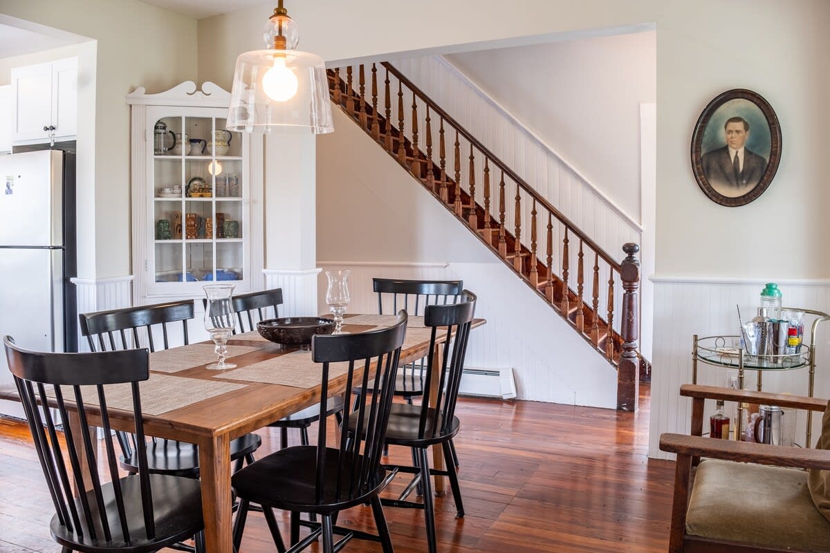 Dining area featuring a wooden dining table with six black spindle-back chairs, set on dark hardwood floors. A prominent wooden staircase with dark banisters leads upstairs, and a white hutch displays dishware.