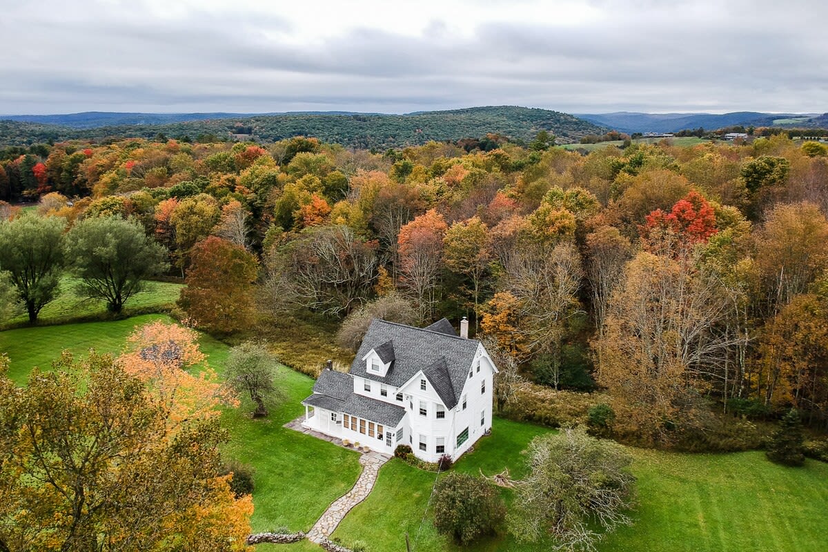 High aerial view showcasing a white two-story house with a dark roof, surrounded by expansive green lawns and a dense forest displaying vibrant fall foliage, with rolling hills in the distant background under an overcast sky.