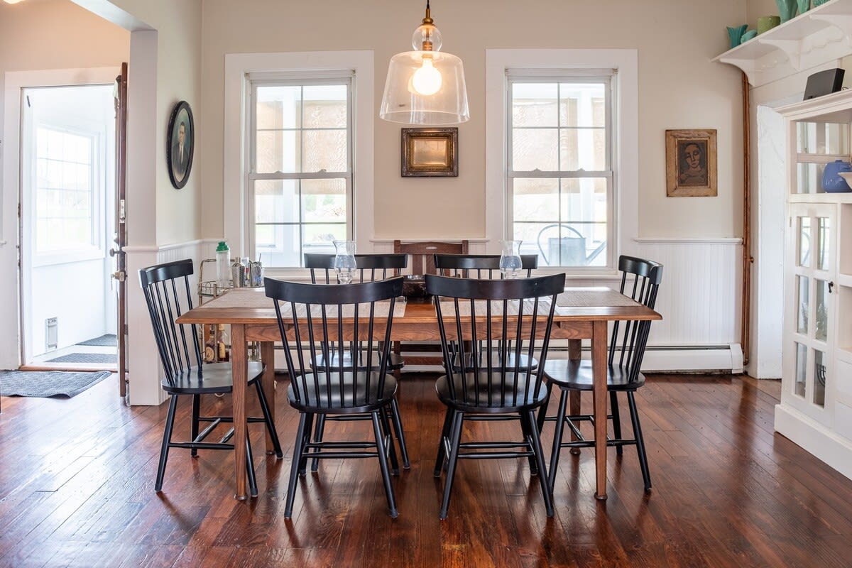 A dining room with light walls and dark hardwood floors features a rectangular wooden table surrounded by six black Windsor-style chairs. Two large windows flank a framed picture on the wall, and a clear glass pendant light hangs above the table.
