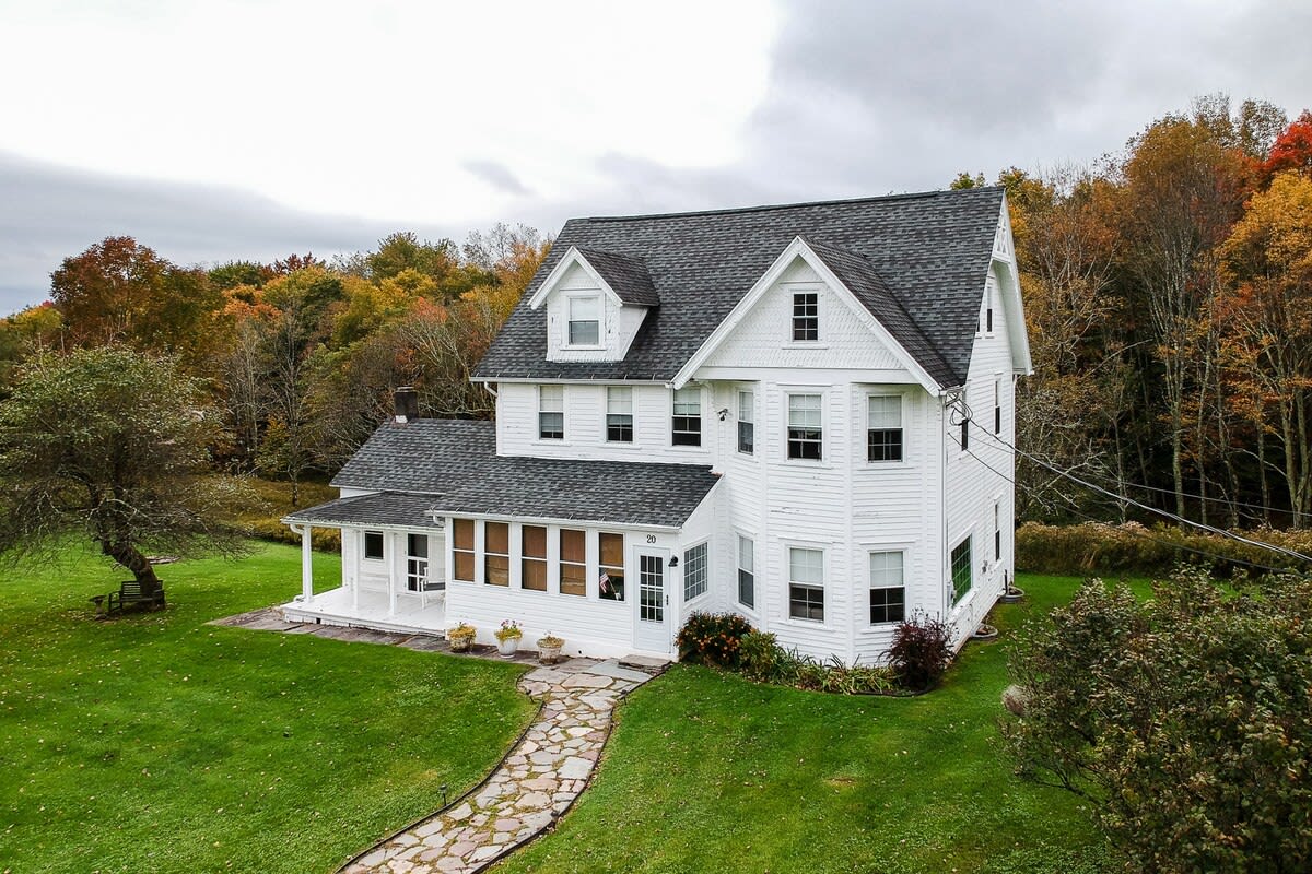 A charming white two-story house with a dark gray gabled roof and multiple dormer windows, featuring a wrap-around porch and a stone pathway leading to the front door. The house is surrounded by a well-maintained green lawn and autumn trees.