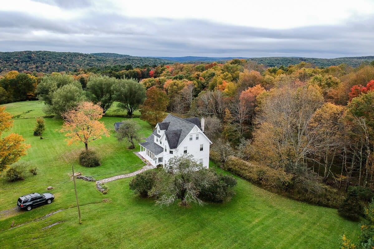 An elevated aerial view of a white multi-story house with a dark roof, situated on a large green property surrounded by rolling hills and a vibrant autumn forest. A dark SUV is parked on the left side of the lawn.