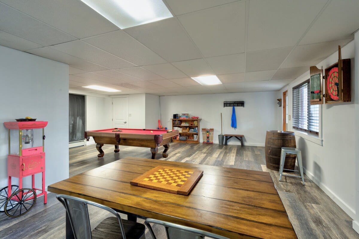 Spacious game room with laminate flooring, featuring a pool table, a wooden table with a chessboard, a popcorn machine, and a dartboard, under a drop ceiling with fluorescent lighting.