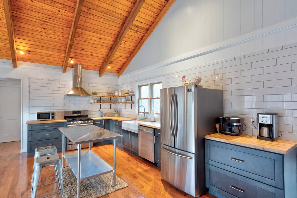 Wide shot of the kitchen showcasing dark gray cabinetry, light wood countertops, a large stainless steel refrigerator, and a central stainless steel island with bar stools. The vaulted wood ceiling and white subway tile backsplash add to the bright, modern farmhouse aesthetic.