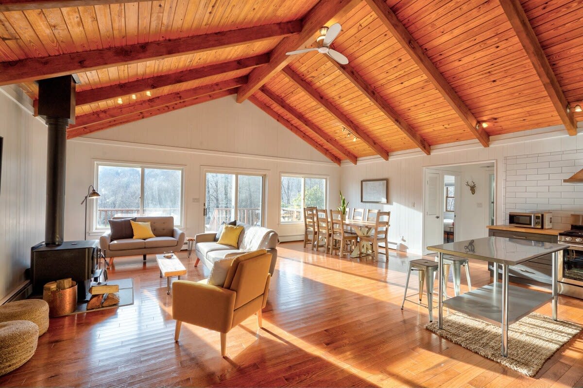 Bright, open-concept living area with a vaulted wooden ceiling, hardwood floors, a wood-burning stove, and large windows offering scenic views, connecting to a dining area and a modern kitchen.