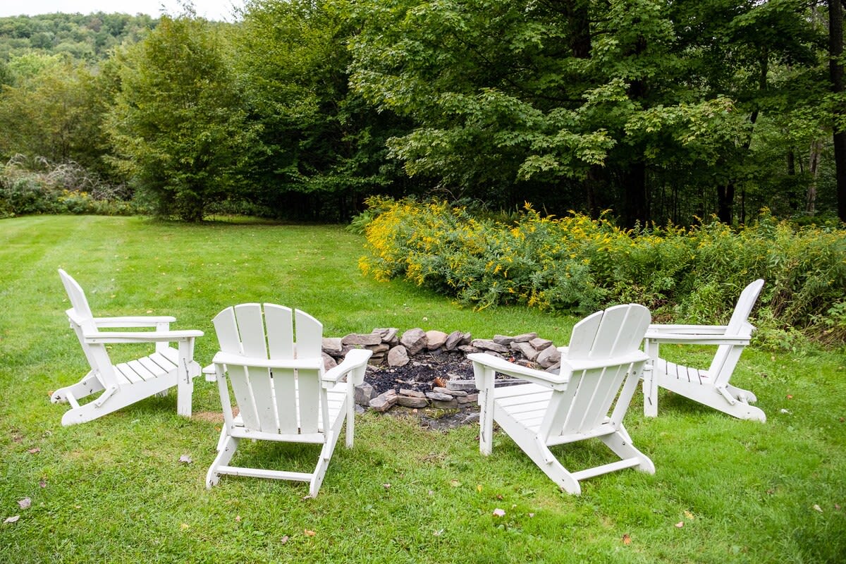 A cozy outdoor fire pit area is set on a vibrant green lawn, featuring a stone-ringed fire pit surrounded by four white Adirondack chairs. Lush trees and yellow wildflowers frame the background.