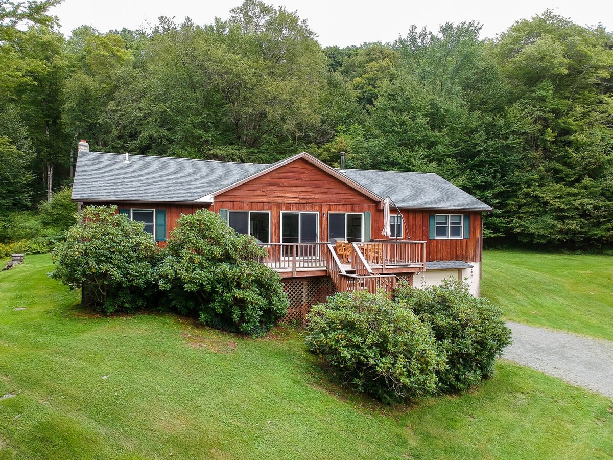 Aerial view of a rustic red-sided house with a gray roof, surrounded by lush green grass and dense forest, featuring a wooden deck with outdoor seating.