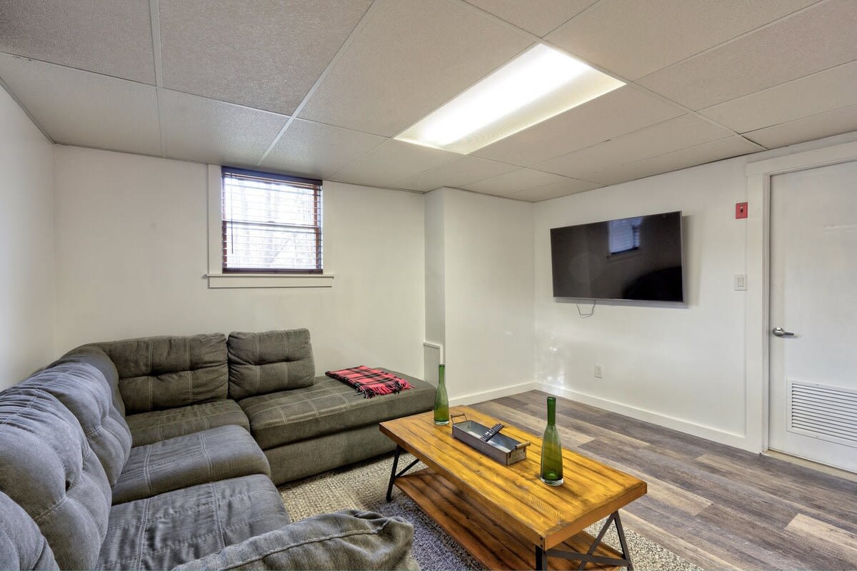 Cozy media room with a large gray sectional sofa, a wooden coffee table, and a wall-mounted TV, featuring a drop ceiling and a small window with blinds.