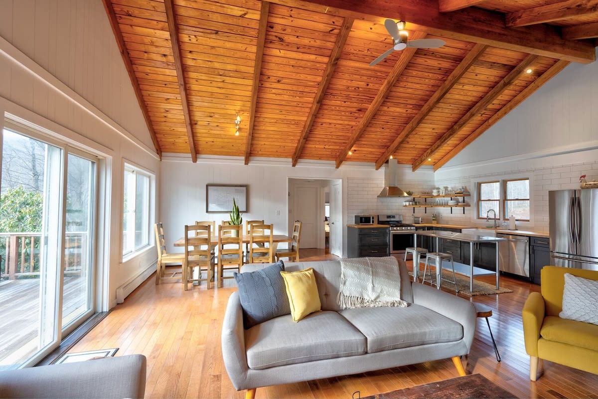 Wide shot of an open-concept living space featuring a vaulted wooden ceiling, hardwood floors, a gray sofa, a yellow armchair, a dining area with a wooden table, and a modern kitchen with dark cabinets and a stainless steel island.