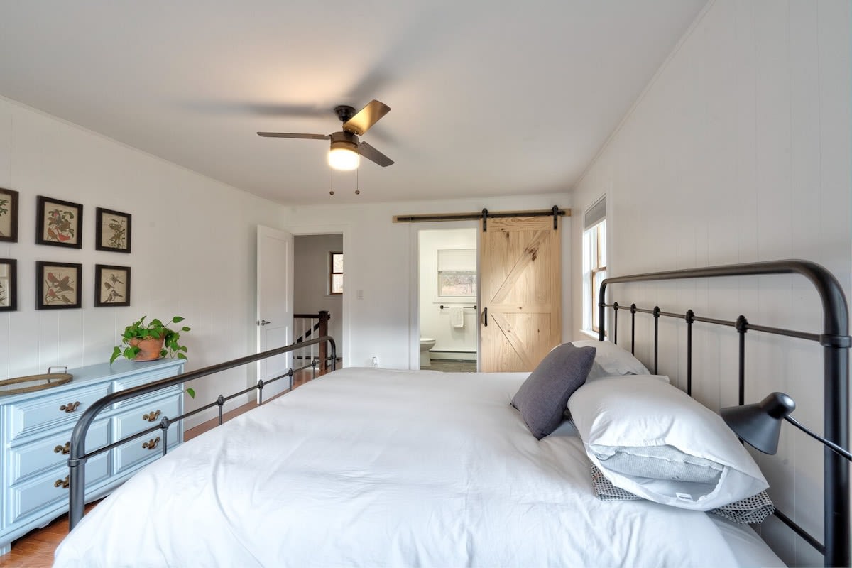 Another angle of bedroom 1, showing the black metal bed frame with white bedding, a light blue dresser, and a rustic wooden barn door leading to an en-suite bathroom. White walls and hardwood floors create a clean, inviting space.