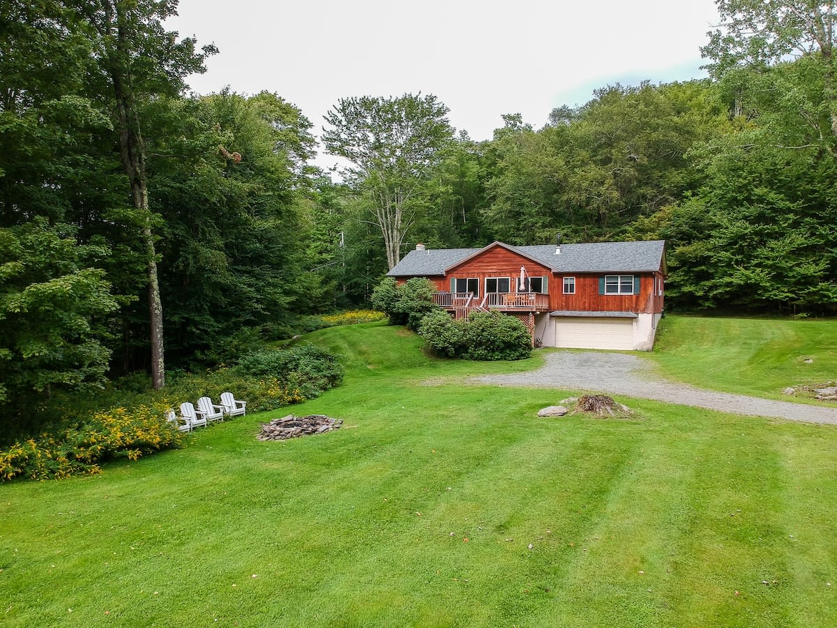 An aerial view showcases a rustic wooden chalet nestled amidst a lush green landscape with dense trees. A gravel driveway leads to the house, and a fire pit area with white chairs is visible on the expansive lawn.
