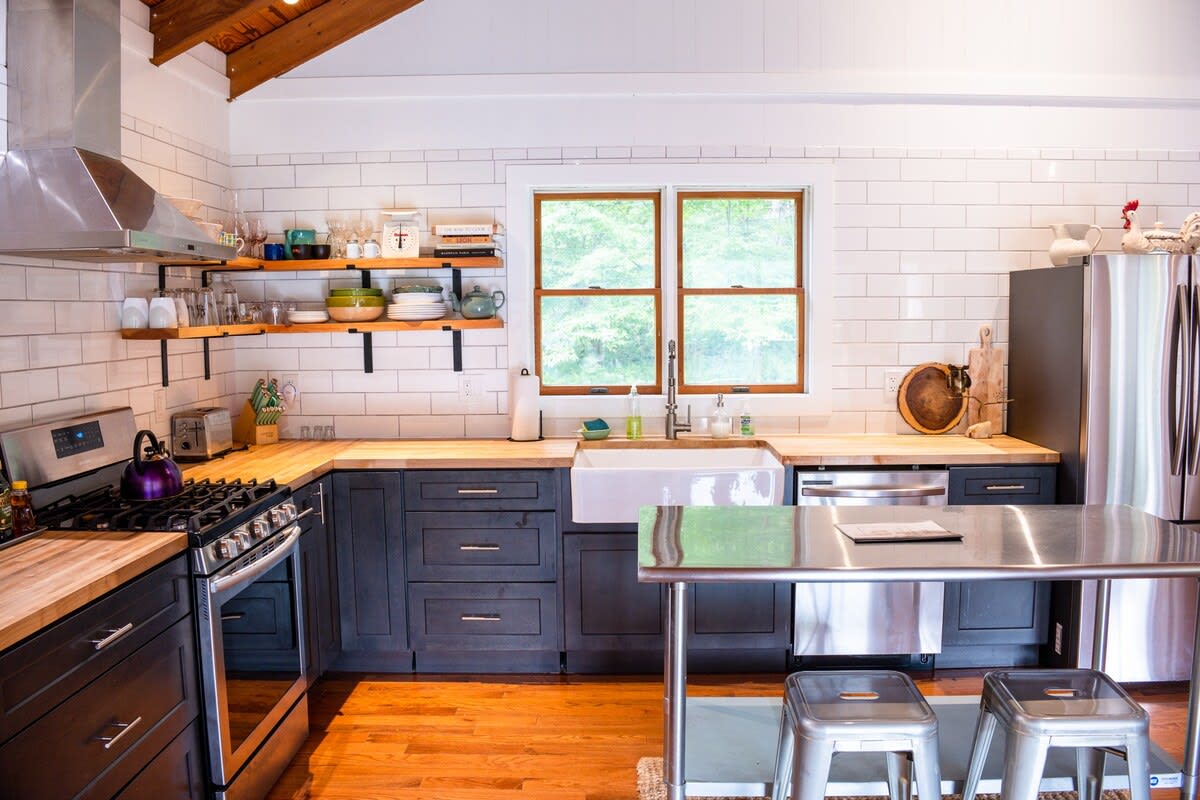Kitchen view highlighting the white farmhouse sink, large window overlooking trees, and open shelving with dishes. Dark gray base cabinets and light wood countertops contrast with the white subway tile and warm hardwood floors.