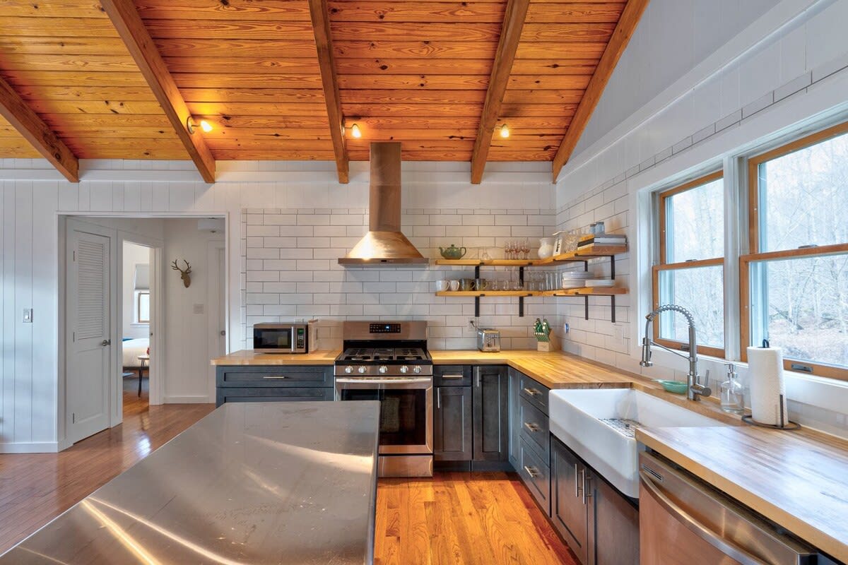 Modern kitchen with dark gray cabinets, butcher block countertops, white subway tile backsplash, and a vaulted wood-paneled ceiling with exposed beams. A stainless steel island and appliances complete the contemporary rustic look.