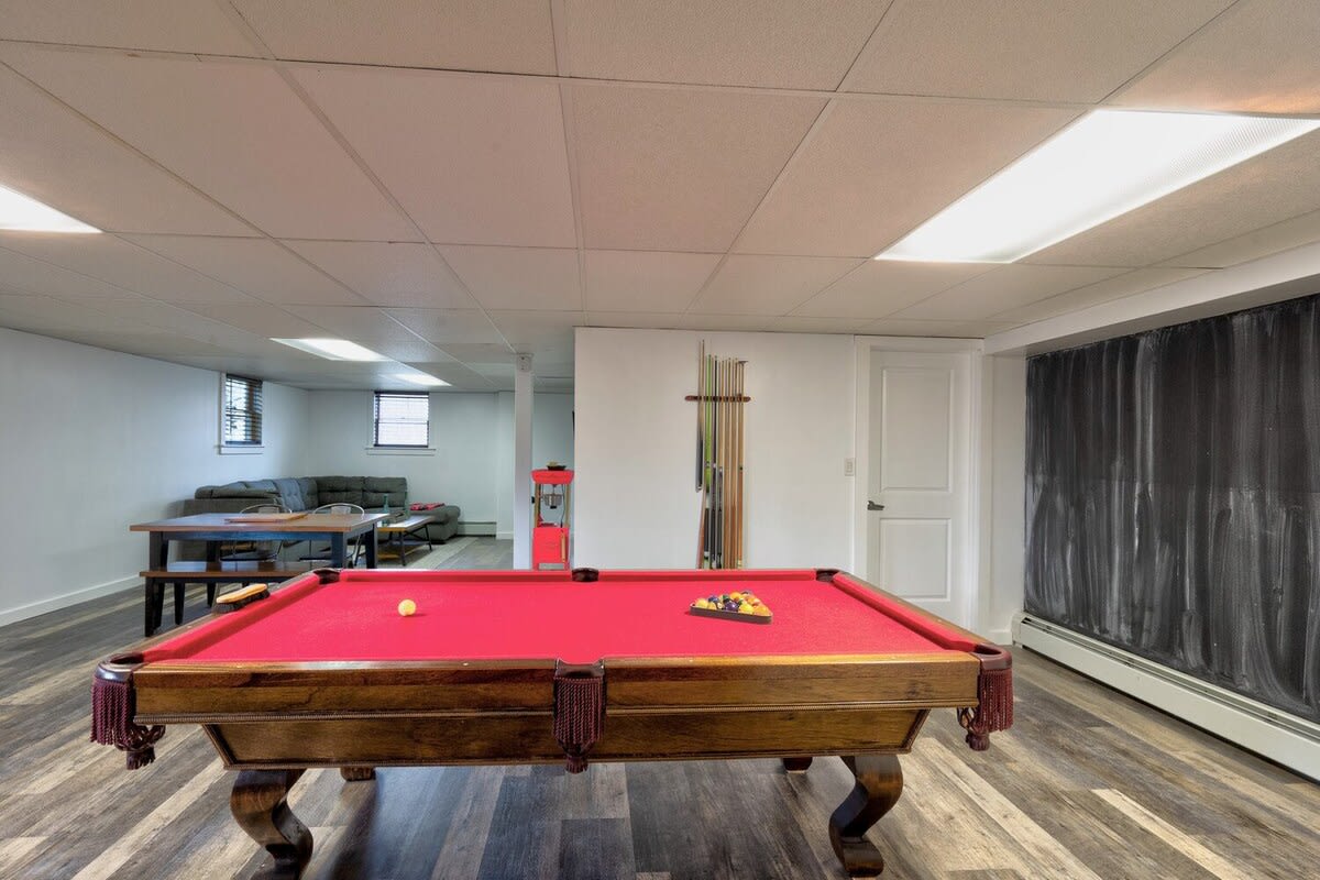A spacious game room features a prominent red-felt pool table with a wooden base, set on dark wood-look laminate flooring. In the background, a casual living area with a sofa and table is visible, all under a drop ceiling.