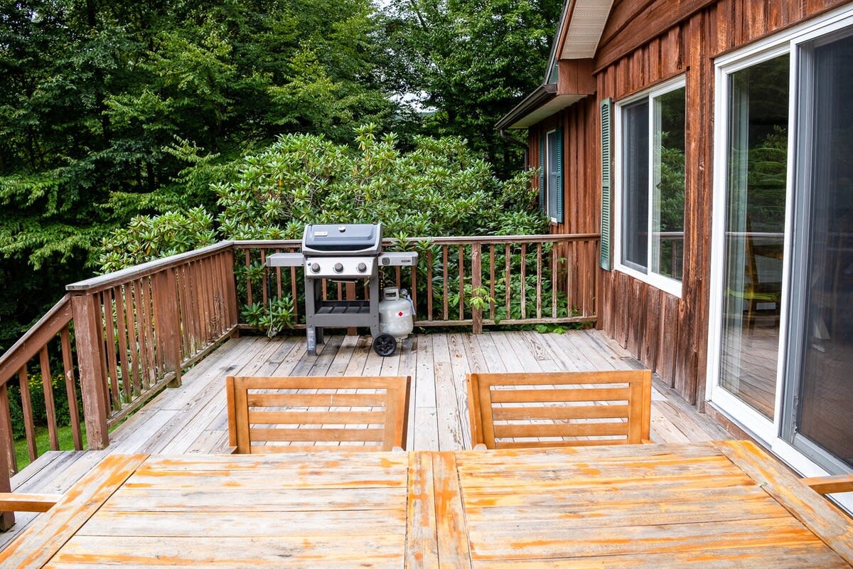 A rustic wooden deck offers an outdoor dining area with a large wooden table and chairs, alongside a gas BBQ grill. The deck is surrounded by lush green trees, providing a natural, private setting.