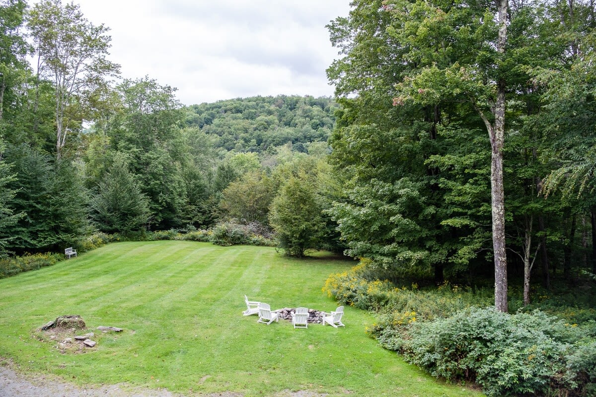 Expansive green lawn surrounded by dense forest and rolling hills, featuring a stone fire pit with four white Adirondack chairs, offering a serene outdoor gathering space.
