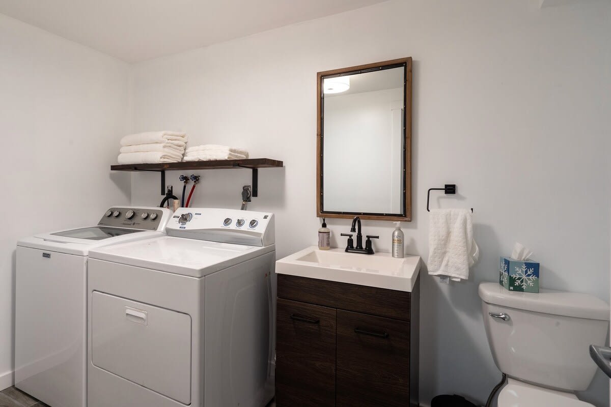 A functional laundry room doubles as a half bathroom, featuring a white washer and dryer, a dark wood vanity with a white sink, and a toilet. A wooden-framed mirror hangs above the sink, and a shelf holds folded towels.