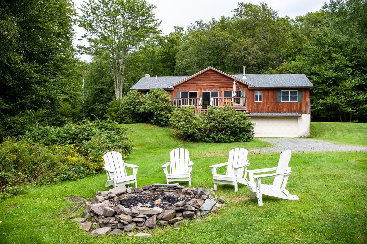 Rustic wooden chalet nestled in a lush green landscape with mature trees, featuring a large deck, a gravel driveway leading to a garage, and a fire pit area with white Adirondack chairs on the lawn.