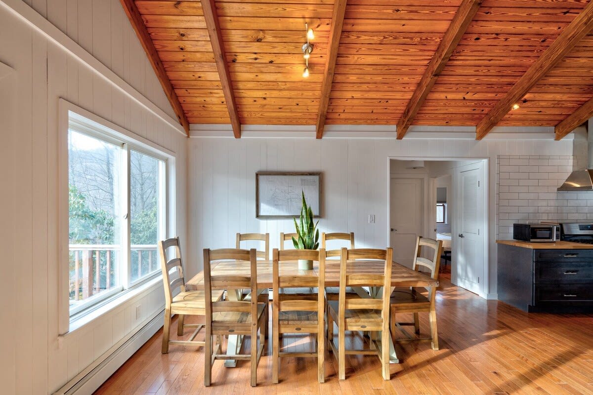 Bright dining area with a large wooden table and matching chairs, set against white vertical shiplap walls and a vaulted wooden ceiling, with a large window overlooking the outdoors.