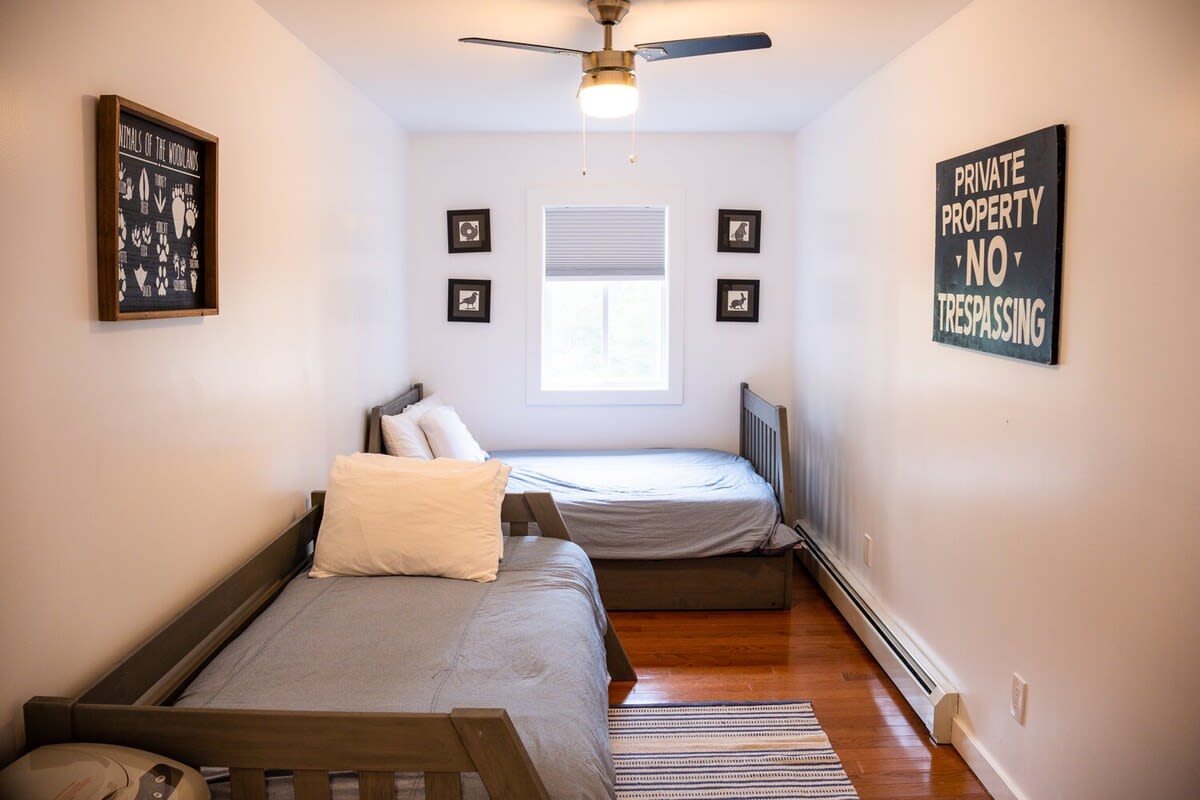 A bright, narrow bedroom features two twin beds with gray bedding, a central window with blinds, and a ceiling fan. White walls are adorned with framed art, and the room has warm hardwood floors.