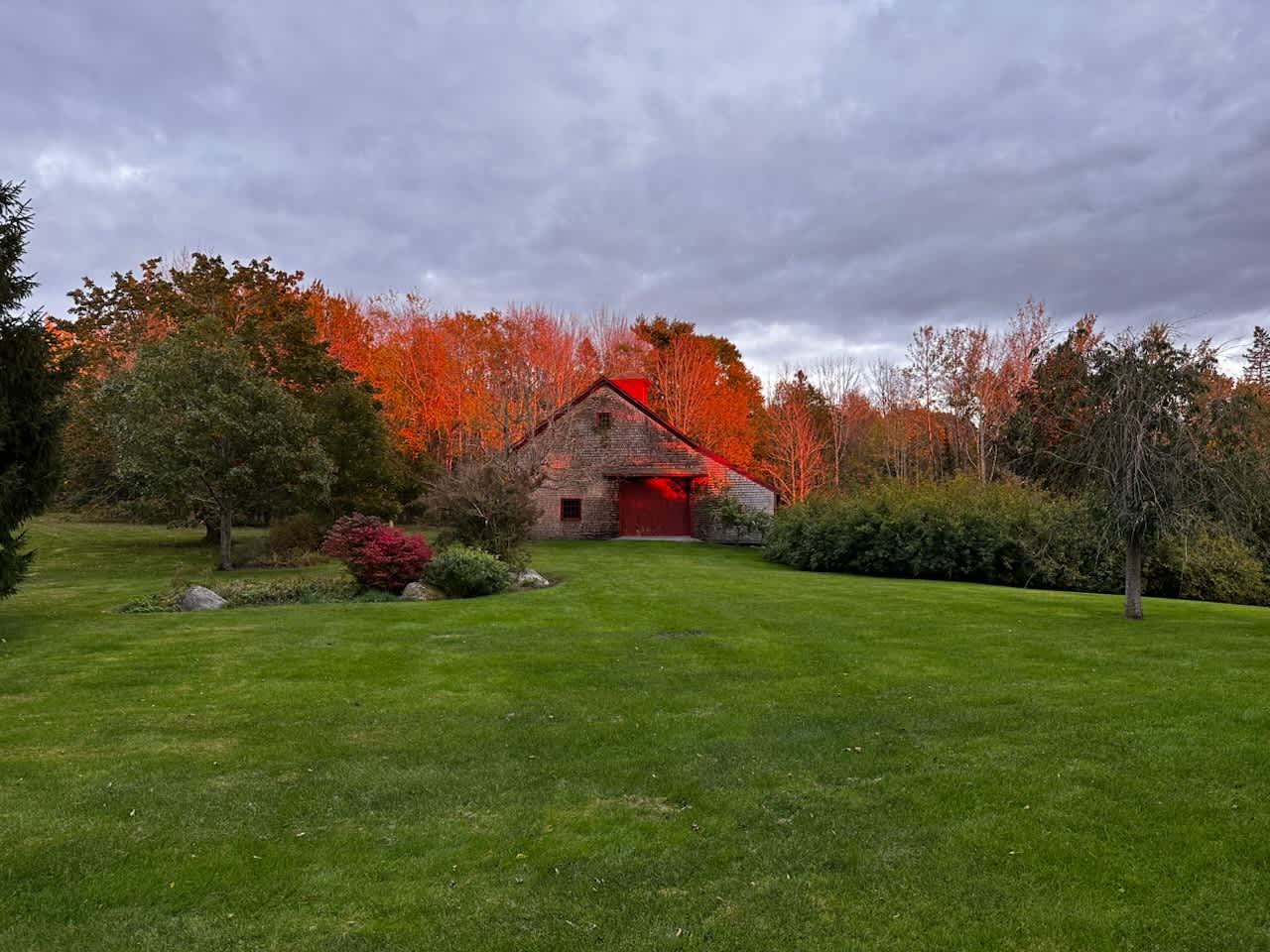 A rustic barn with a red facade and shingled roof, nestled among trees with vibrant orange and red foliage, illuminated by the warm glow of sunset, overlooking a vast green lawn under a cloudy sky.