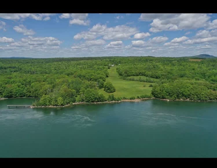 Aerial view showcasing a vast waterfront property with a long dock extending into the teal water, surrounded by lush green forests and a central grassy clearing where a house is nestled under a blue sky with scattered clouds.
