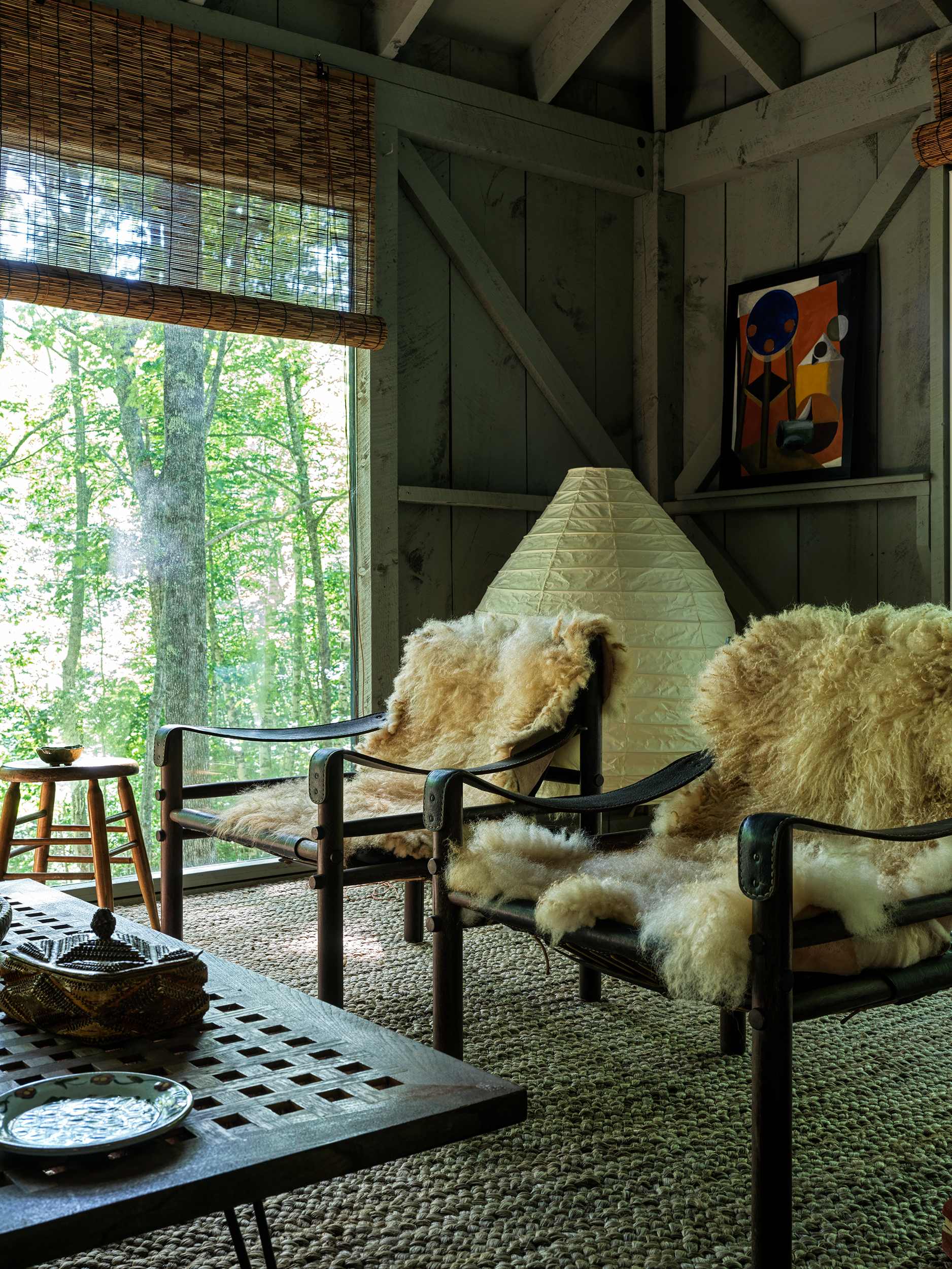 Rustic living room corner with two black chairs draped in sheepskin throws, a tall paper lantern floor lamp, and an abstract artwork on the paneled wall, all illuminated by natural light from large windows overlooking the forest.