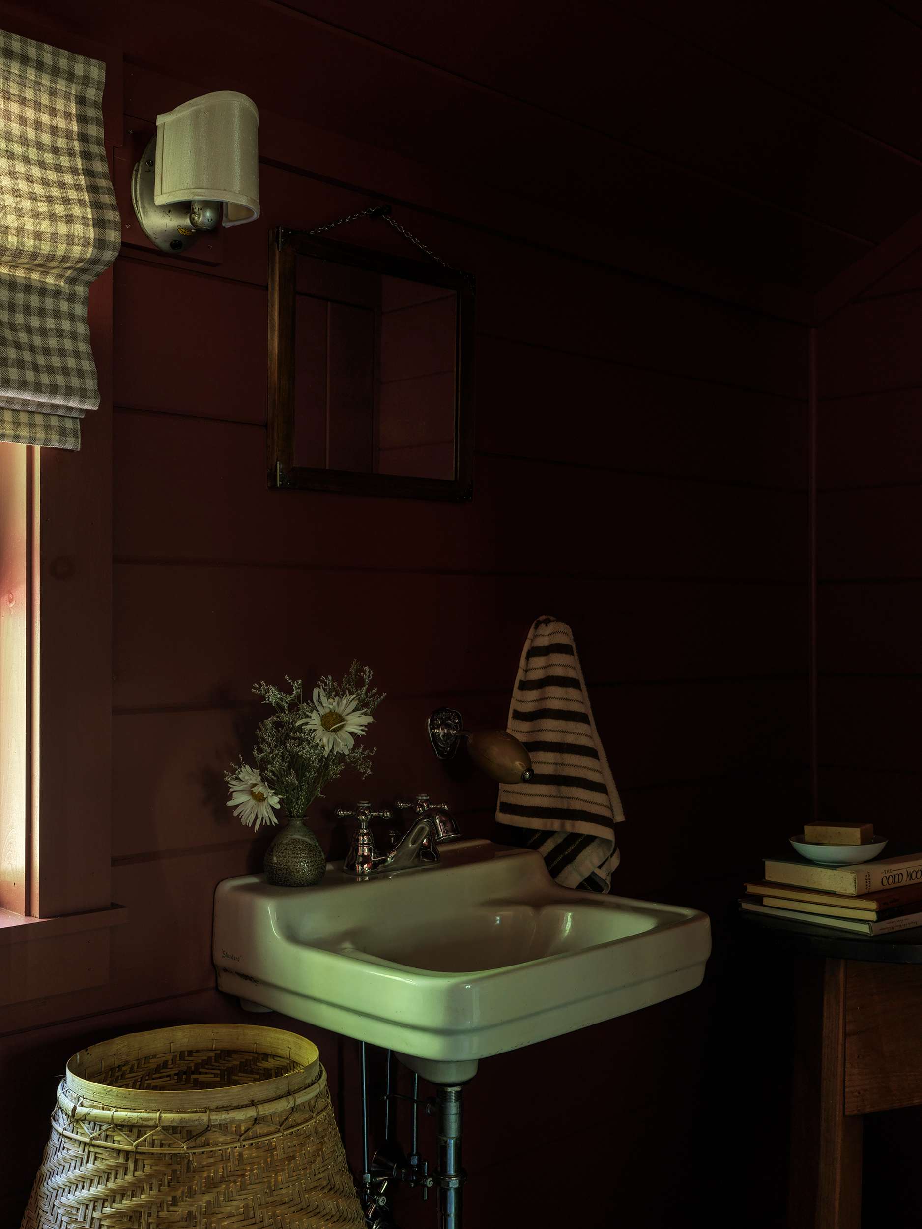 Compact bathroom featuring dark red wooden plank walls, a classic white pedestal sink with chrome fixtures, a small mirror, and a window with a plaid roller blind, creating a cozy and rustic feel.