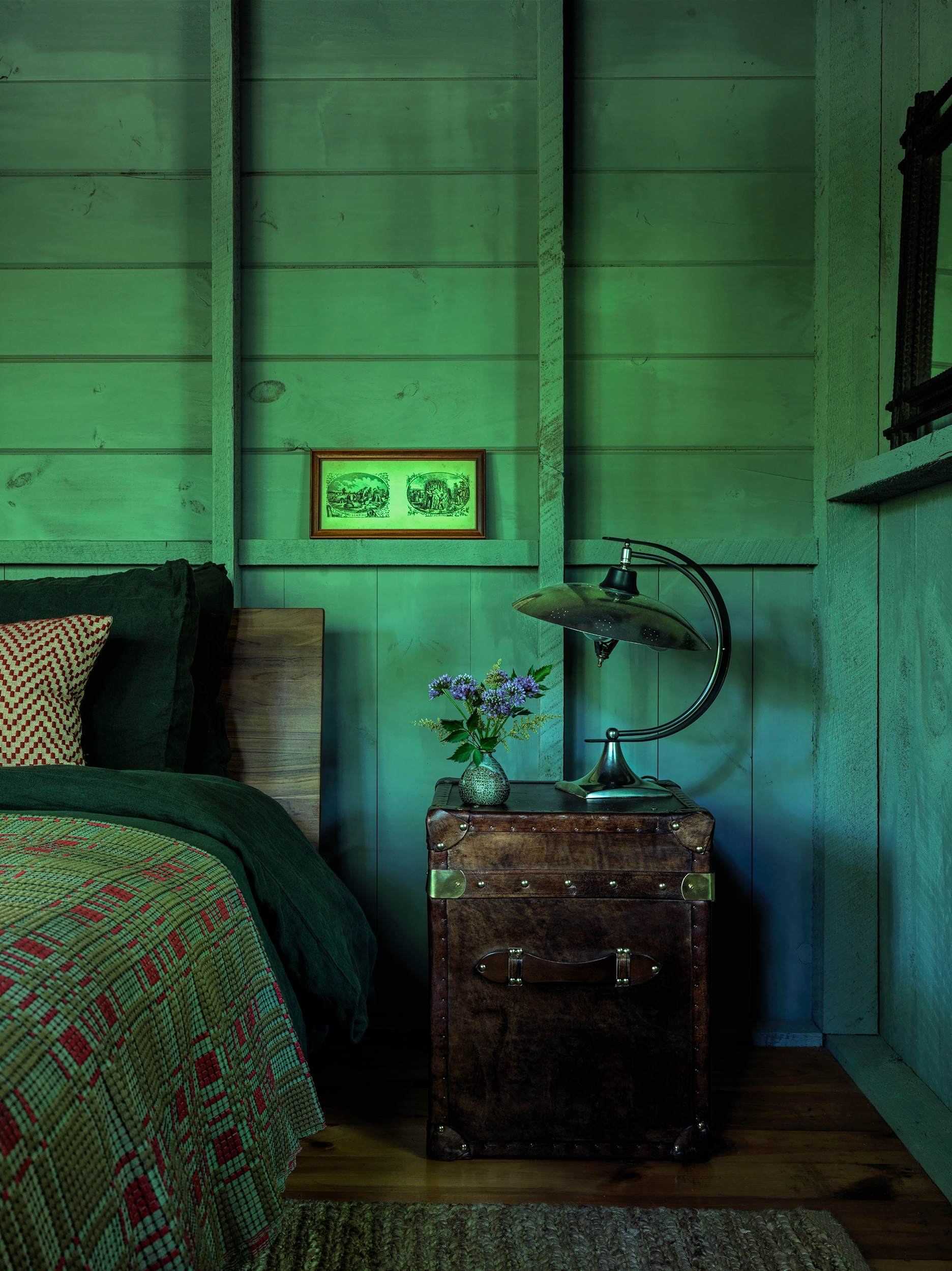 Serene bedroom with green-painted wooden plank walls and exposed studs, featuring a wooden bed with dark green bedding, a vintage trunk nightstand, and a distinctive metal lamp, creating a rustic and cozy ambiance.