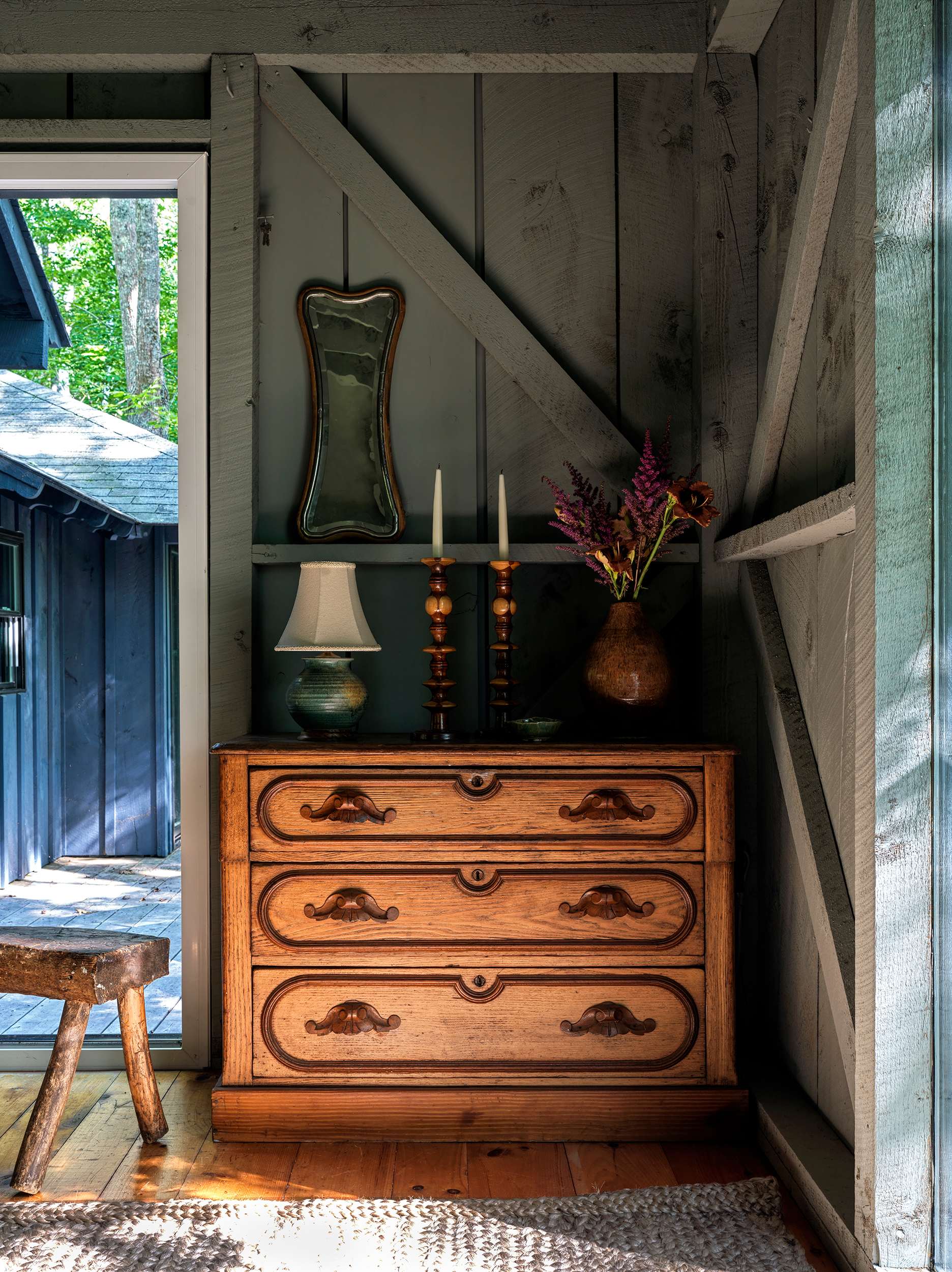 Inviting entryway featuring a carved wooden dresser adorned with a lamp, candles, and a vase of flowers, beneath a decorative mirror, with an open door revealing an outdoor path and another building.