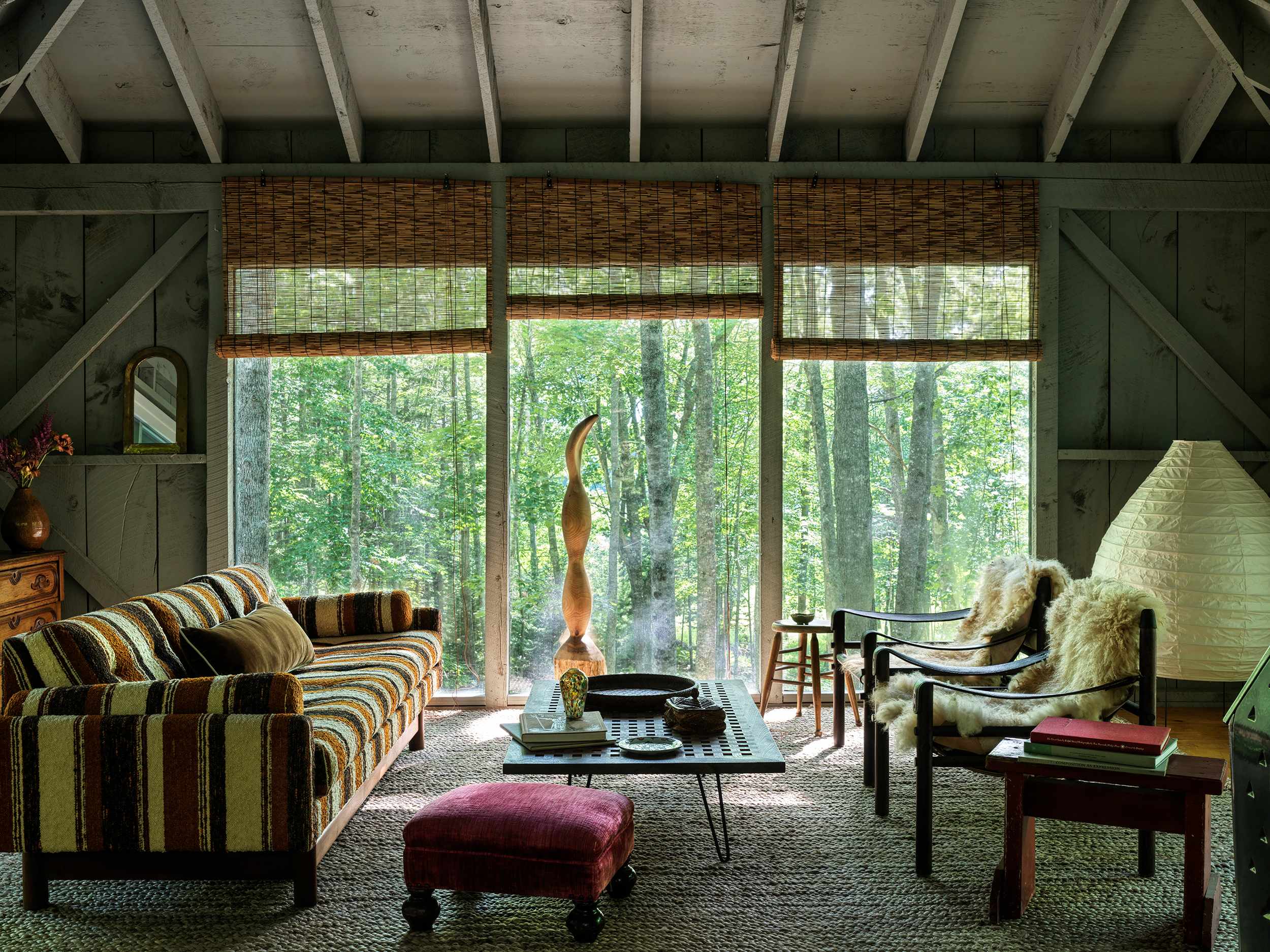 Cozy living room featuring a striped sofa, two black chairs with sheepskin throws, and a wooden coffee table on a woven rug, all set against large windows offering immersive forest views. The room has exposed wooden beams and rustic paneling.