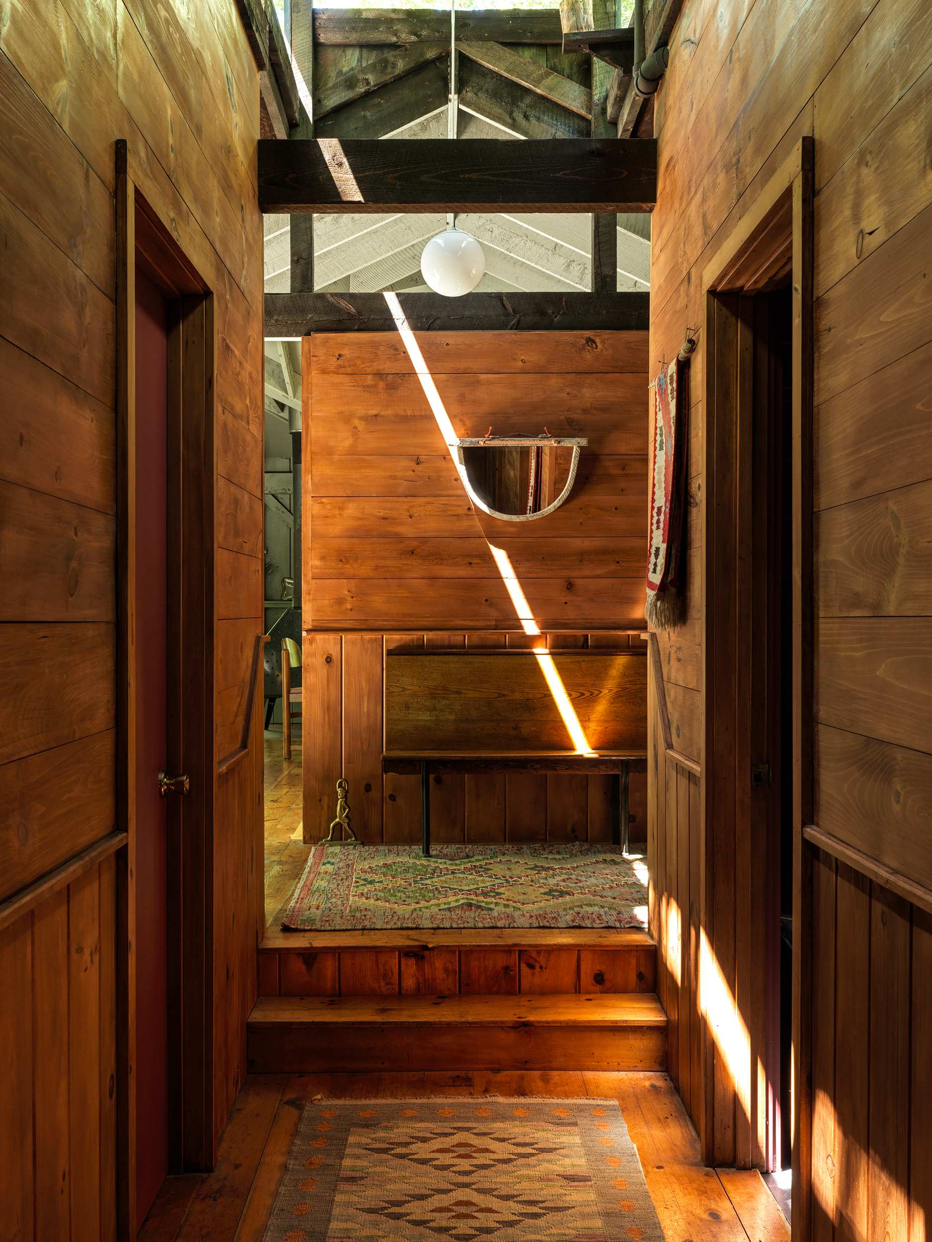Warm wooden hallway with paneled walls and floors, featuring a short staircase leading up, exposed ceiling beams, and two doorways on either side, with natural light streaming in.