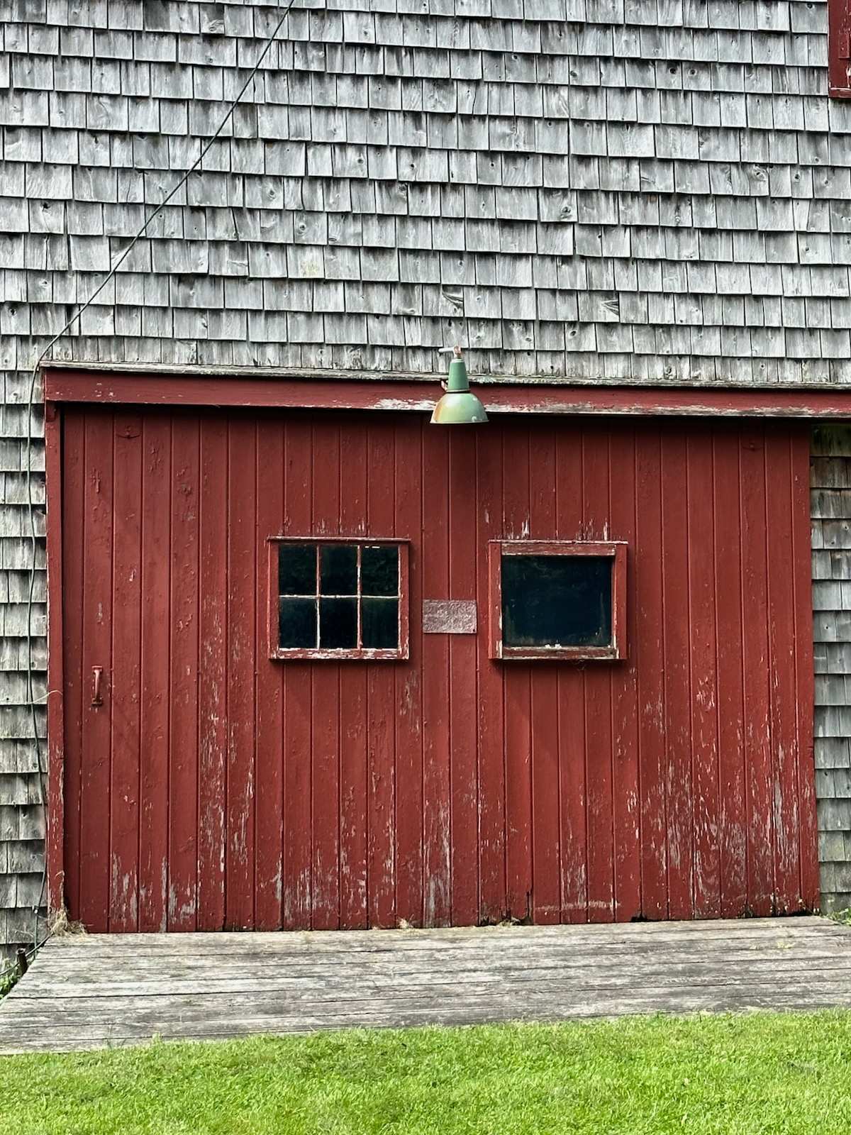 A close-up of a rustic red barn wall with weathered wooden planks, featuring two small windows with dark panes and a vintage green industrial light fixture hanging above, all beneath a shingled roof.