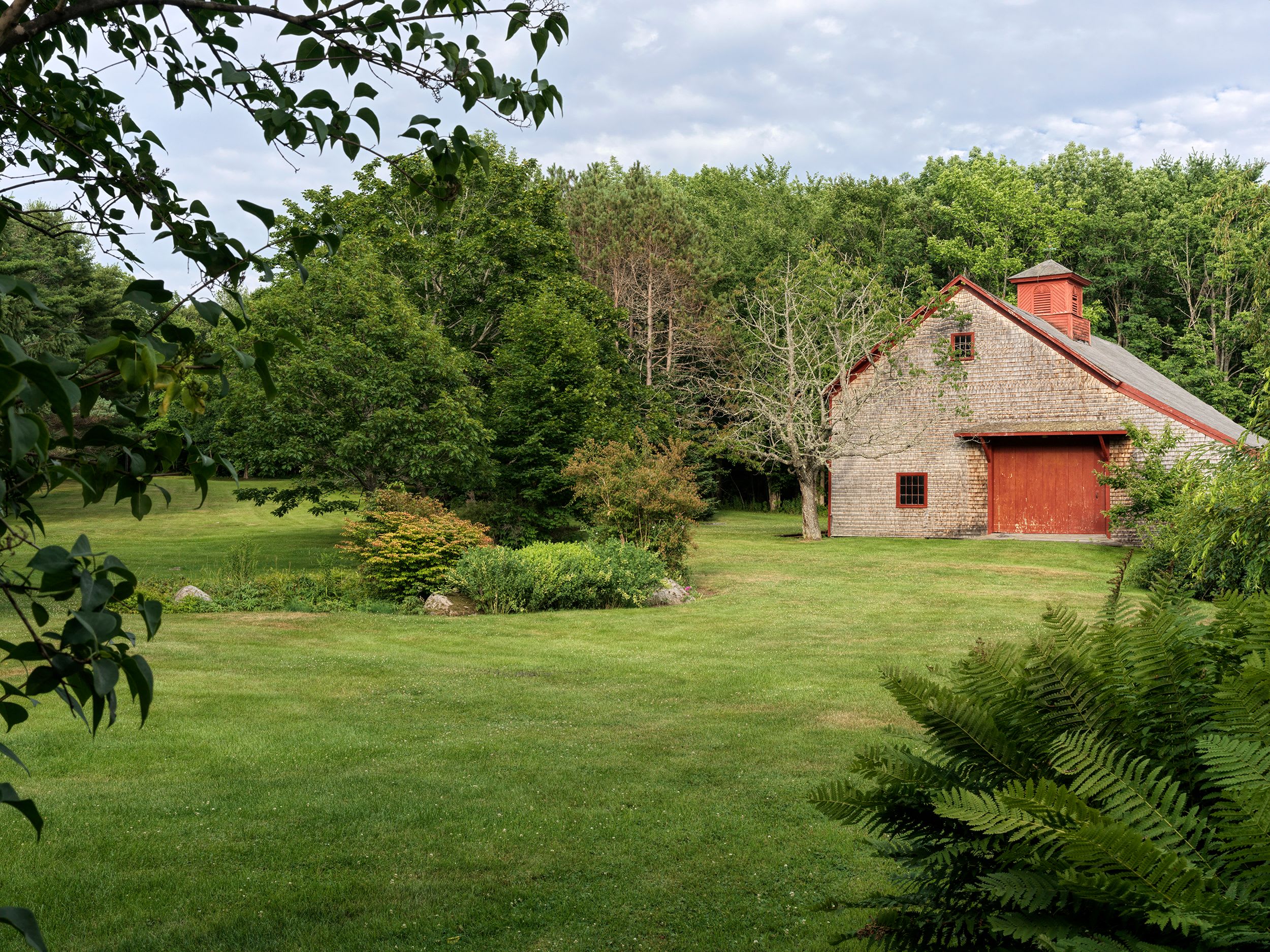A rustic shingled barn with a vibrant red door and a cupola stands prominently in a vast, manicured green lawn. Lush trees frame the property under a bright, clear sky, with a small garden bed in the foreground.