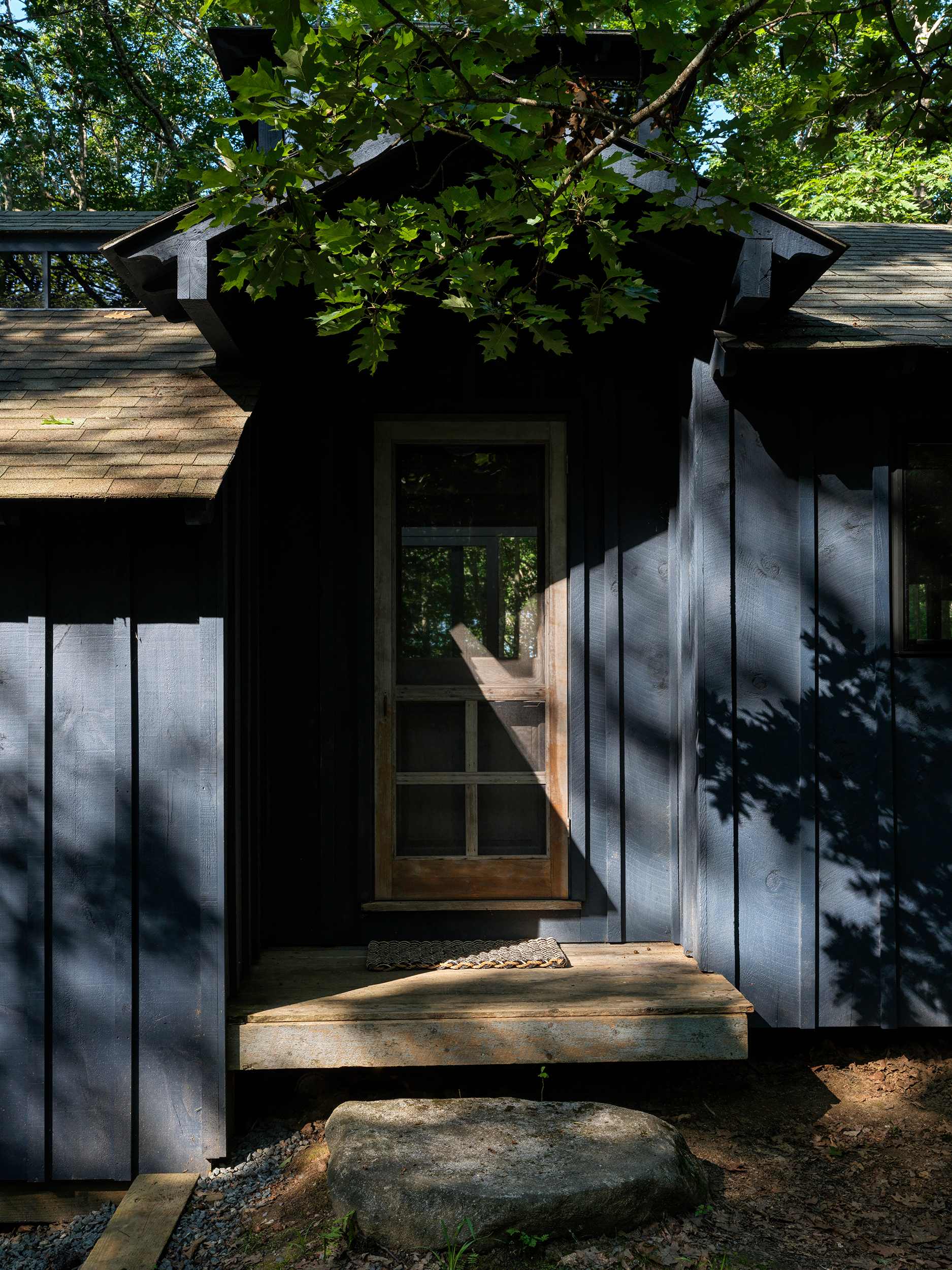 The front entrance of a dark blue wooden cabin, featuring a screen door with glass panels and a small wooden porch, partially illuminated by sunlight filtering through overhead trees.